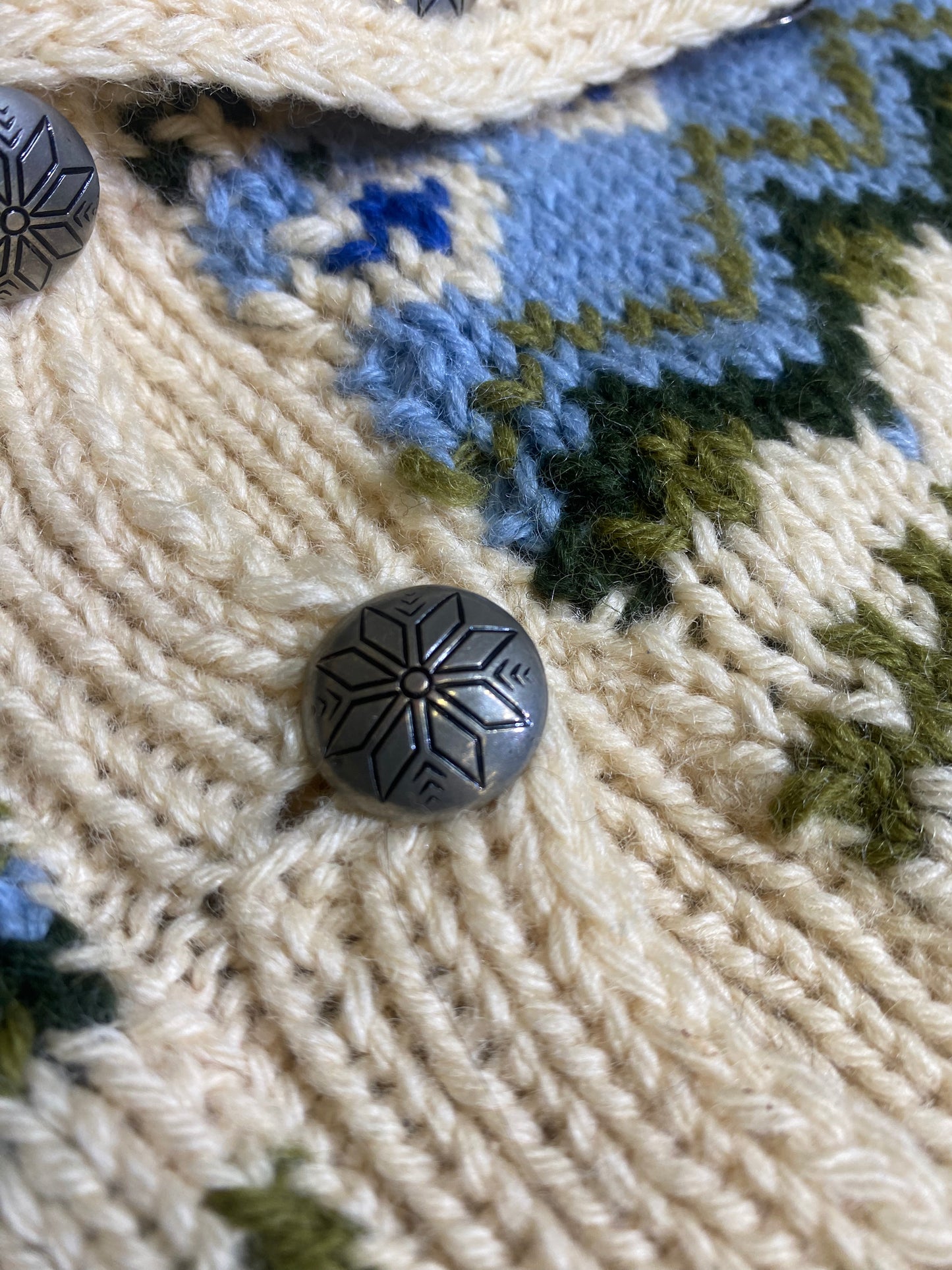 Close-up image of a metal button on the Vintage Hand Knit Norwegian Cardigan Sweater. The button features an elegant engraved snowflake pattern, adding a touch of sophistication and vintage charm to the sweater's front closure.